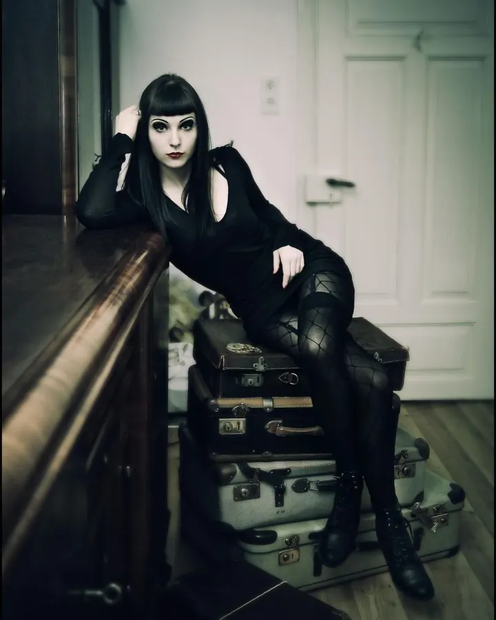Femme trans latino aux cheveux noirs et frange, en robe noire ajustée, collants à mailles et bottines, assise sur des valises anciennes dans un décor sombre.