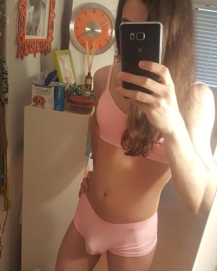 Femme trans européenne aux cheveux bruns, prenant un selfie dans un miroir, en sous-vêtements roses, dans une salle de bain avec horloge orange et cadre décoratif.