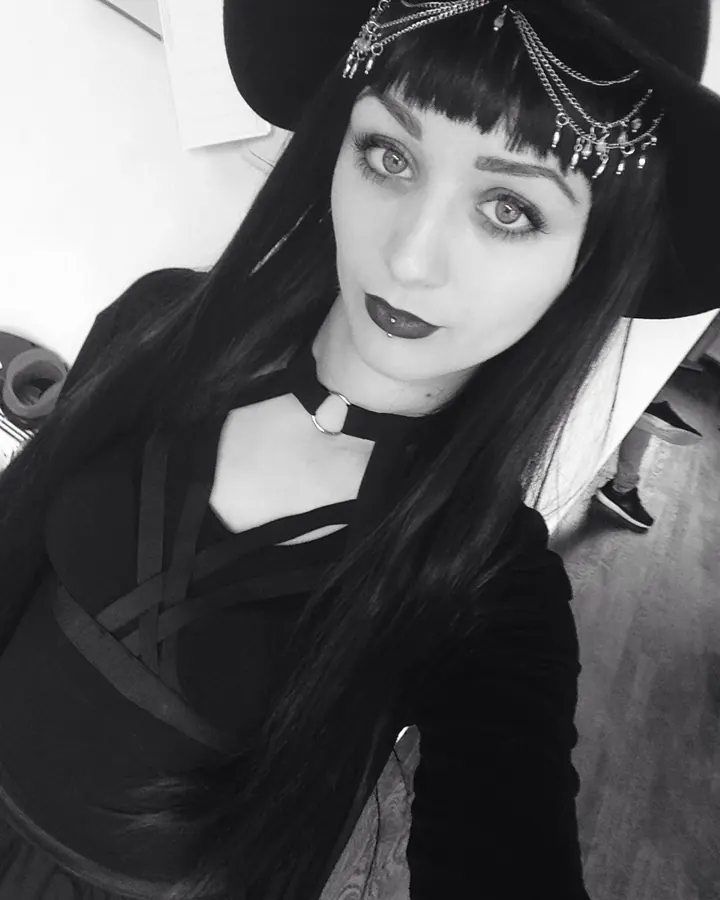 Femme trans aux cheveux noirs, portant un chapeau noir et un haut noir, regardant l'objectif dans un cadre intérieur en noir et blanc.