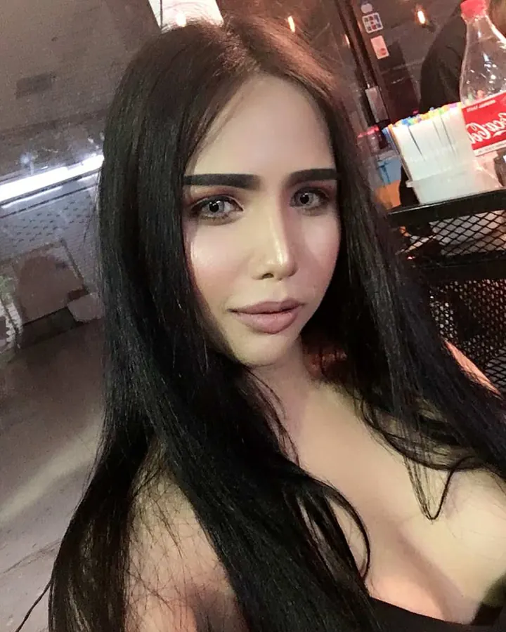 Femme trans latine aux cheveux bruns-noirs, en haut blanc, posant devant un fond rose
