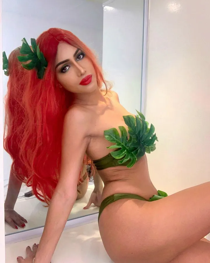 Femme trans aux cheveux roux, regardant l'appareil photo, portant un collier fin et une tenue sombre, dans un intérieur sobre.