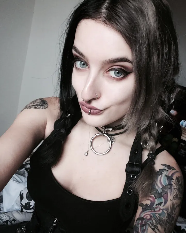 Femme trans aux cheveux bruns-noirs, regardant l'appareil photo, portant un collier fin et une tenue sombre, dans un intérieur sobre.