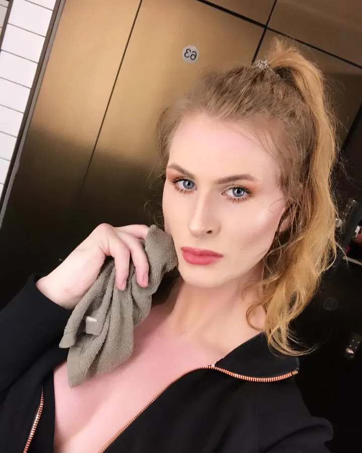 Femme trans en tenue noire, cheveux blonds, regardant l'appareil dans un ascenseur
