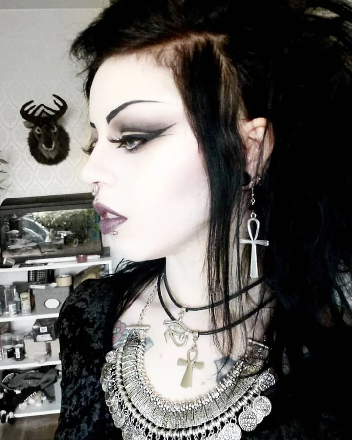 Femme trans aux cheveux noirs longs, portant un haut noir, posant sur un lit, regardant l'appareil avec un sourire.