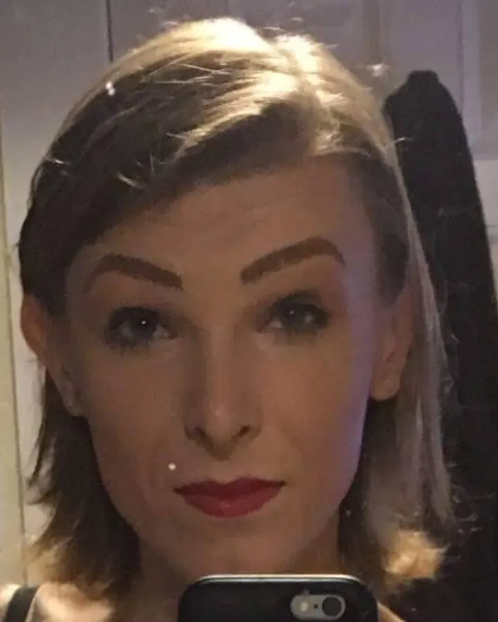 Femme trans aux cheveux blonds courts, regardant l'appareil photo, maquillage subtil, lèvres rouges, dans un intérieur.