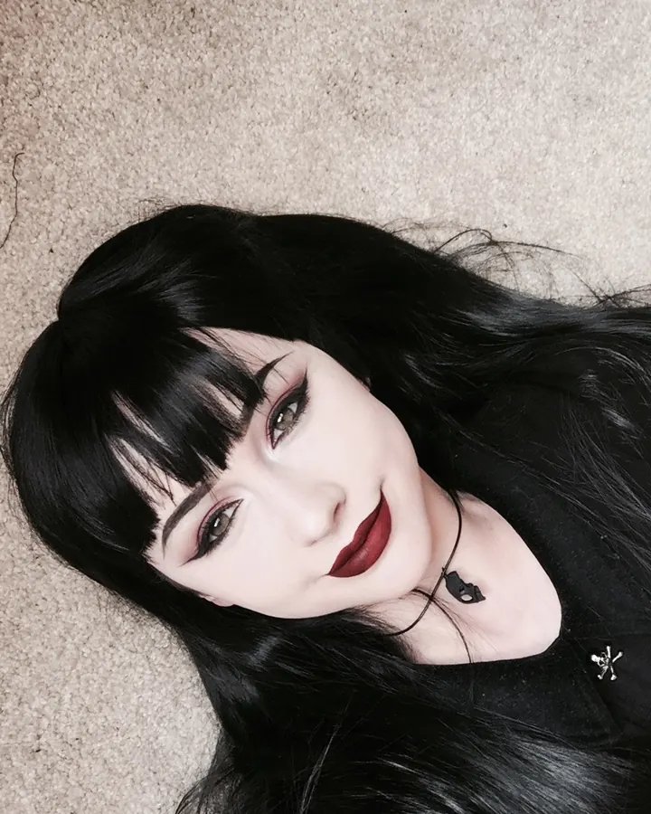 Femme trans allongée sur un tapis, cheveux noirs longs, maquillage sombre, regard vers le haut, collier noir avec pendentif
