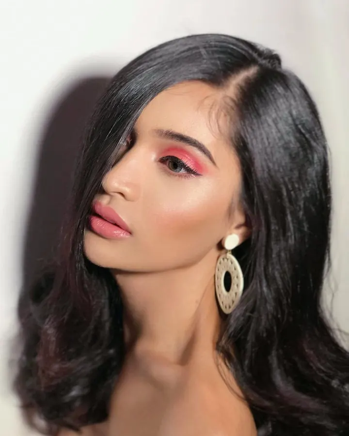 Femme trans aux cheveux bruns-noirs et yeux gris, maquillée avec des ombres à paupières roses, portant de grandes boucles d'oreilles blanches.