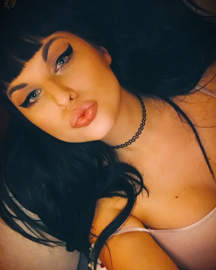 Femme trans asiatique aux cheveux bruns-noirs et yeux bleus, en lingerie noire, allongée sur un lit, avec une lumière douce en arrière-plan.