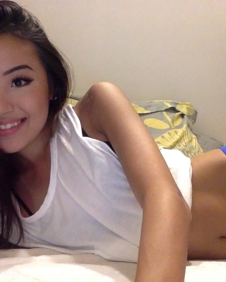 Femme trans asiatique aux cheveux bruns-noirs et yeux noirs, souriante, allongée sur un lit blanc, portant un débardeur blanc et un string bleu.