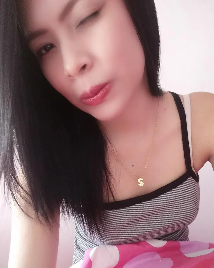 Femme trans aux cheveux noirs longs, portant un haut rayé noir et blanc, avec un collier dollar, regardant vers le bas avec une expression douce.