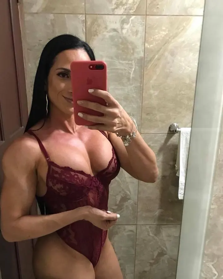 Femme trans aux cheveux noirs longs en body rouge dentelle, prenant un selfie dans une salle de bain, montrant son corps musclé.