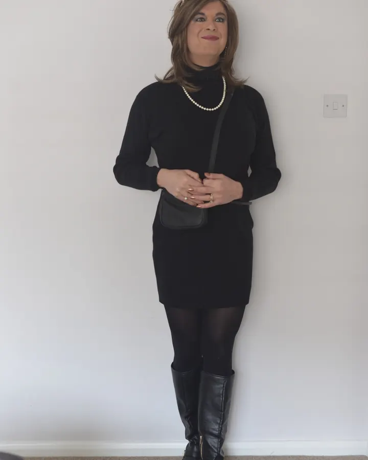 Femme debout contre un mur blanc, vêtue d'une robe noire à col roulé, de collants noirs et de bottes hautes, portant une perle et des lunettes de soleil sur la tête.