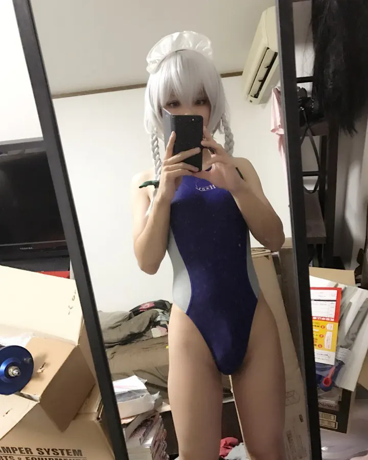 Femme en maillot de bain bleu nuit et blanc, coiffée d'une perruque blanche avec un bonnet de servante, prenant un selfie devant un miroir dans une pièce encombrée.