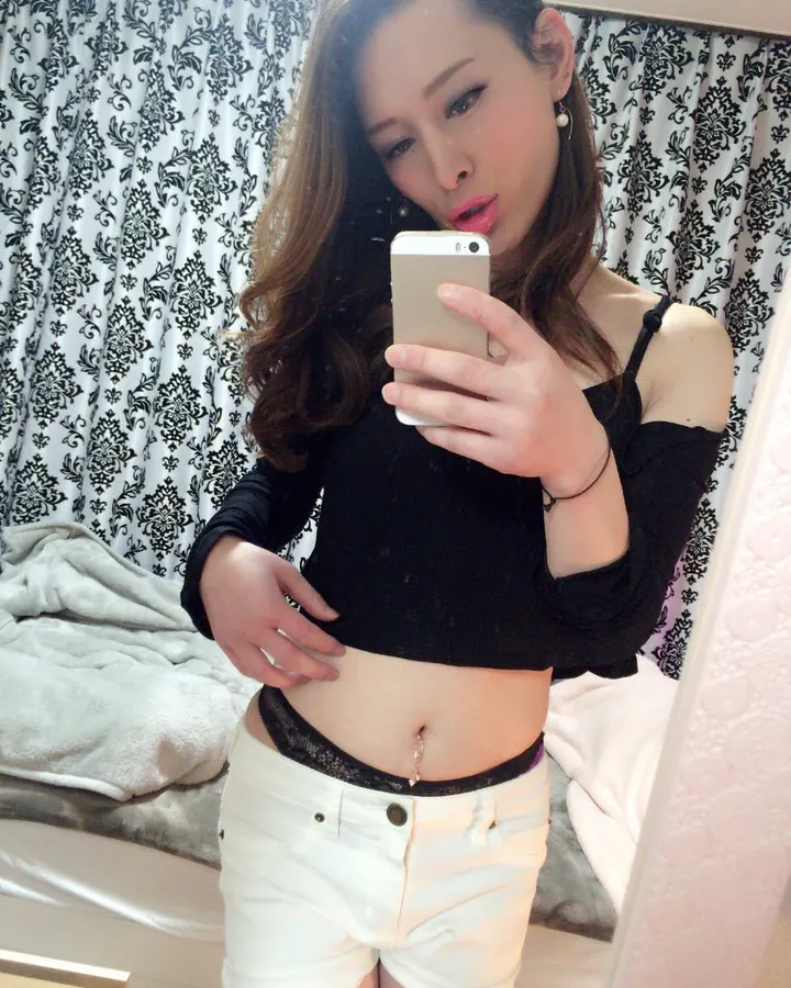 Femme trans aux cheveux châtains, prenant un selfie, portant un haut noir.