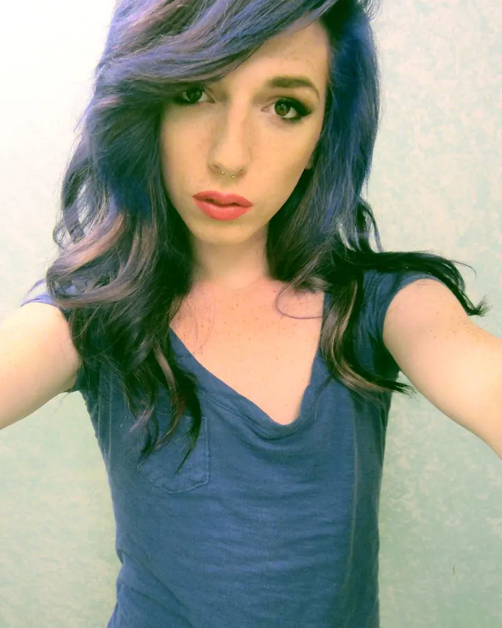 Femme trans aux cheveux bleu foncé ondulés, peau claire avec taches de rousseur, portant un débardeur bleu marine, regard fixe vers l'appareil.