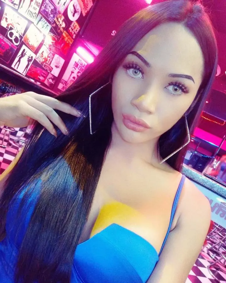 Femme trans aux cheveux noirs longs, maquillage discret, portant un haut noir et un bas noir, posant sur un lit avec un string perlé.