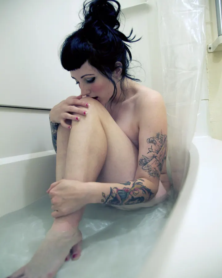Femme trans aux cheveux bruns mi-longs, maquillage naturel, portant un haut noir et un bas noir, posant sur un lit.